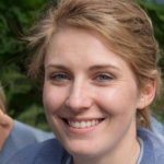Smiling person with short, wavy, light brown hair, wearing a blue shirt. Green foliage is visible in the blurred background.