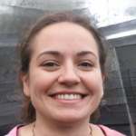 A person with long hair smiling at the camera. Background includes metal and tarp.