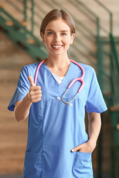Woman in blue medical scrubs with a pink stethoscope smiles and gives a thumbs-up, standing indoors with one hand in her pocket.