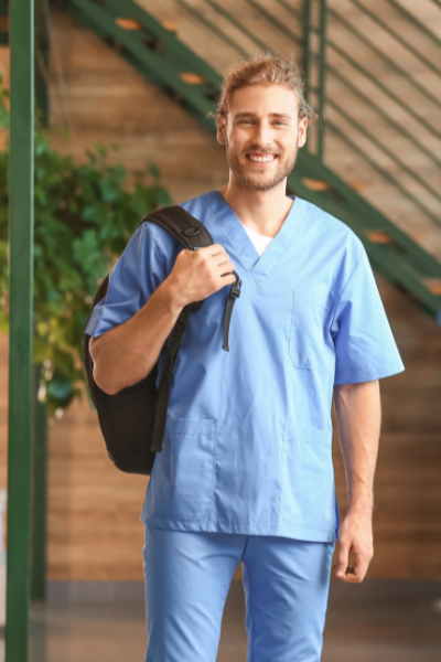 A man wearing light blue scrubs and carrying a black backpack stands indoors, smiling at the camera.